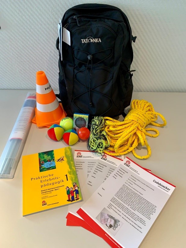 Der Rucksack enthält Übungsmaterialien wie Augenbinden, Jonglierbälle, Spielseil, Stoppuhr, Luftballons, Zauberstab und Stifte, sowie ein Handbuch mit Übungsideen.