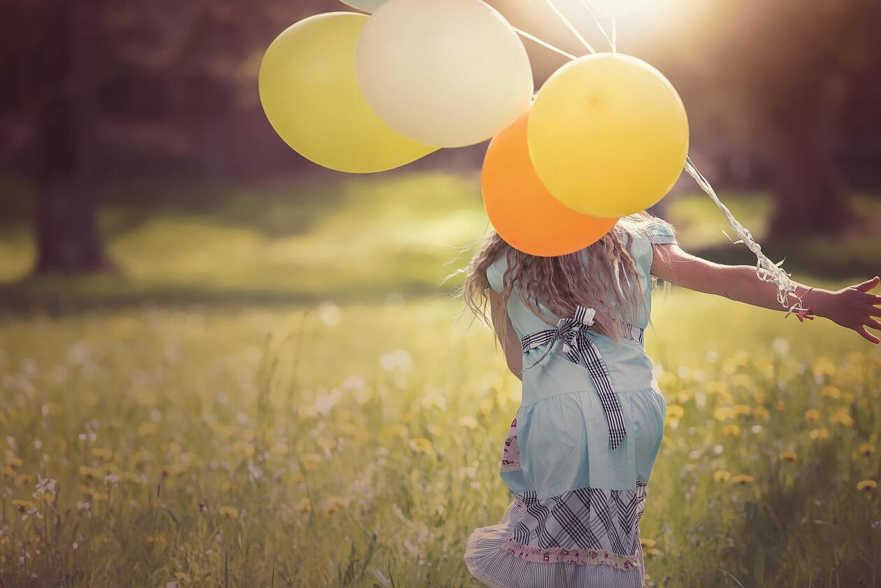 ein Mädchen das Luftballons trägt, läuft durch eine Sommerwiese. Aufnahme von hinten